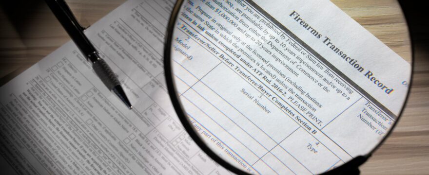 A black pen and a magnifying glass rest on a printed firearms transaction record on a light brown wood table.