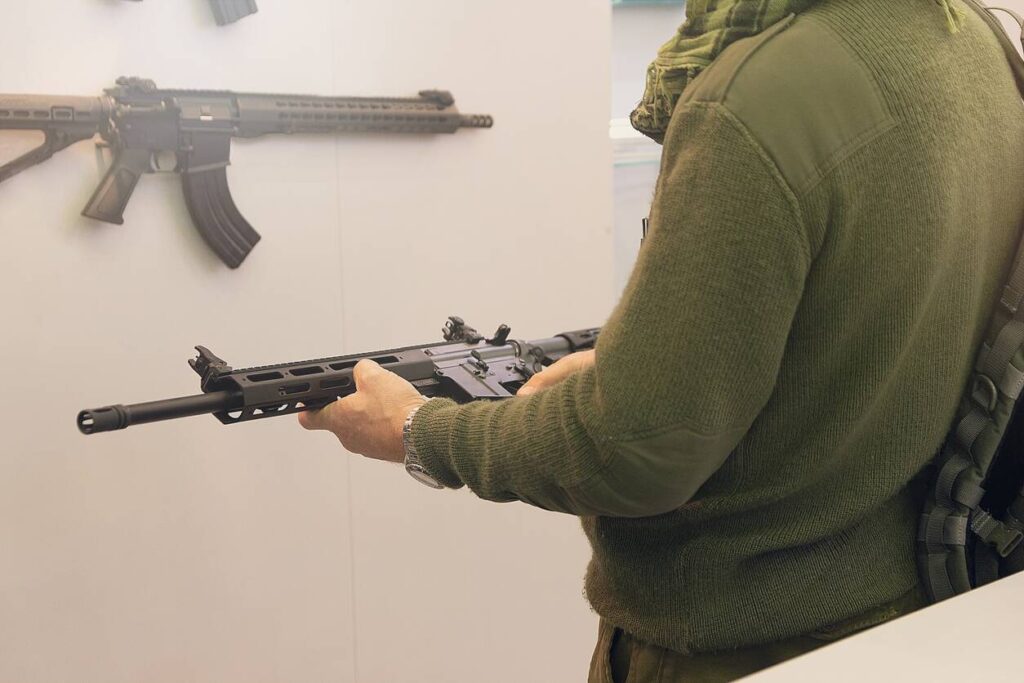 A man wearing a green sweater holds a firearm while another firearm hangs on the display wall in front of him.